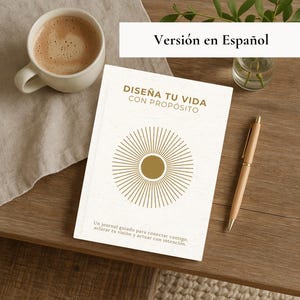 May include: A white journal with gold text that reads "DISEÑA TU VIDA CON PROPÓSITO" and a sunburst design. A cup of coffee, a pen, and a small vase with greenery sit on a wooden table. The journal is a guide to connect with yourself, clarify your vision, and act with intention.