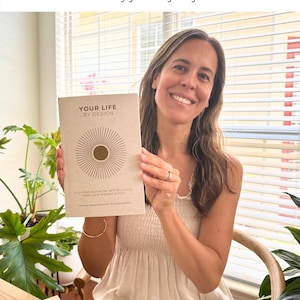 May include: A woman holding a journal titled "YOUR LIFE BY DESIGN." The journal has a beige cover with a sunburst design. The text "Handcrafted with love, from my journey to yours" is above the journal. The journal is for self-discovery, vision, and aligned action.
