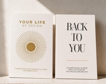 May include: Two off-white guided journals. The left journal features a gold sunburst graphic and the text 'YOUR LIFE BY DESIGN A guided journal for self-discovery, vision, and aligned action.' The right journal reads 'BACK TO YOU A guided space to trade who you should be for who you actually are.'