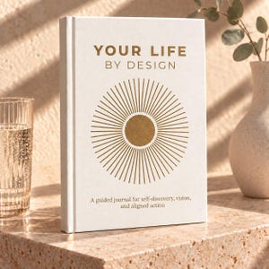 May include: A white hardcover journal titled YOUR LIFE BY DESIGN in gold lettering. The cover features a central gold sunburst graphic and the text A guided journal for self-discovery, vision, and aligned action printed in a smaller gold font.