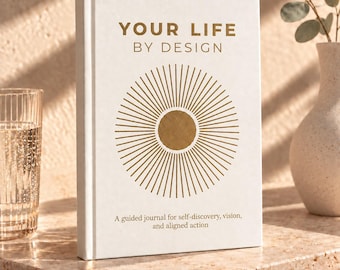 May include: A white hardcover journal titled YOUR LIFE BY DESIGN in gold lettering. The cover features a central gold sunburst graphic and the text A guided journal for self-discovery, vision, and aligned action printed in a smaller gold font.