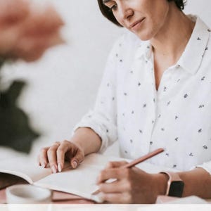 May include: A woman writing in a notebook with a pencil. She wears a white button-down shirt with a small black pattern and a rose gold watch. The text on the image reads: Find clarity. Reconnect with yourself. Move forward with intention.