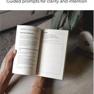 May include: An open book with the title "DEFINE YOUR VISION" and the subtitle "Design Your Aligned Life" is held open. The text is in black on a white page. The book is on a light-colored table. The image is well-lit.