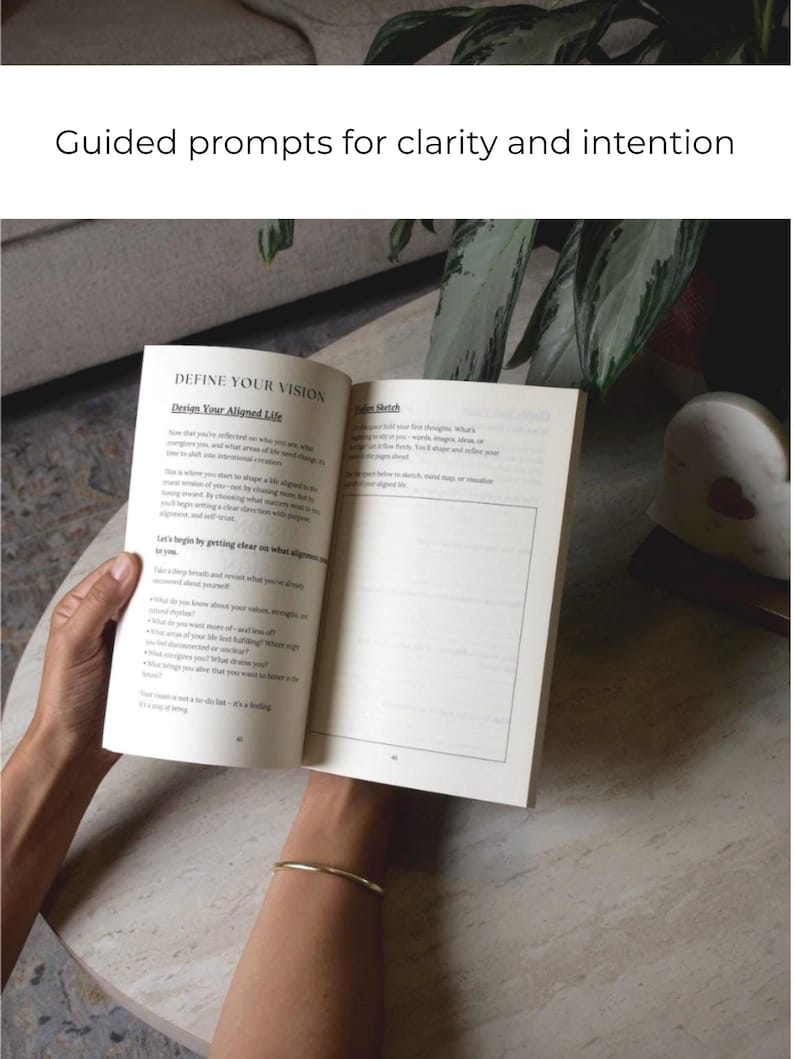 May include: An open book with the title "DEFINE YOUR VISION" and the subtitle "Design Your Aligned Life" is held open. The text is in black on a white page. The book is on a light-colored table. The image is well-lit.