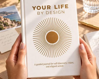 May include: A white hardcover journal titled YOUR LIFE BY DESIGN in gold foil lettering. The cover features a central gold sunburst graphic and the text A guided journal for self-discovery, vision, and aligned action printed in gold.