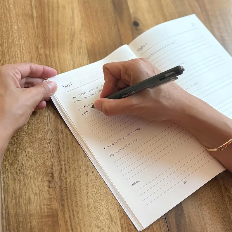 May include: A journal open to a page marked "Day 1", being written on with a black pen. The text "The future belongs to the beauty of their dreams" is visible. A gold bracelet is on the wrist. The journal is on a wooden surface.