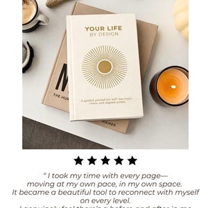 May include: A cream-colored journal titled "YOUR LIFE BY DESIGN" with a gold sunburst design. The journal is on top of a brown book, with a cup of coffee, a candle, and decorative stones nearby. The journal is described as a guided journal for self-discovery.
