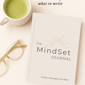 May include: A cream-coloured journal titled "The MindSet Journal" with the text "40 Days to Reprogram Your Mind." A cup of green matcha tea and eyeglasses are next to the journal. The text "You'll always know what to write" is above the journal.