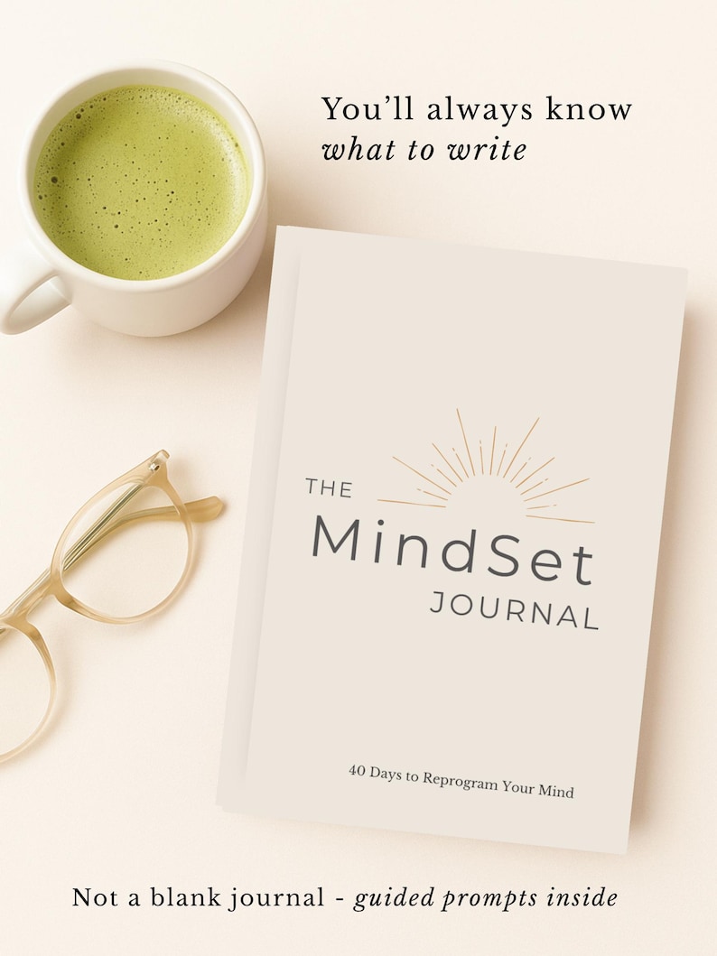 May include: A cream-coloured journal titled "The MindSet Journal" with the text "40 Days to Reprogram Your Mind." A cup of green matcha tea and eyeglasses are next to the journal. The text "You'll always know what to write" is above the journal.