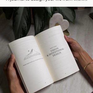 May include: An open journal with the text "A journal to design your life from within." The journal's pages display the text "PART THREE VISION & INTENTIONAL DESIGN" and a mountain illustration. The journal is held open by hands.