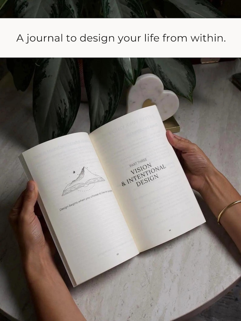 May include: An open journal with the text "A journal to design your life from within." The journal's pages display the text "PART THREE VISION & INTENTIONAL DESIGN" and a mountain illustration. The journal is held open by hands.