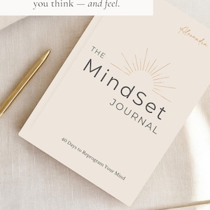 May include: A cream-coloured journal titled "The Mindset Journal" with gold lettering and a sunburst design. The text "40 days that will change the way you think - and feel" is above the journal. A gold pen rests on the surface.