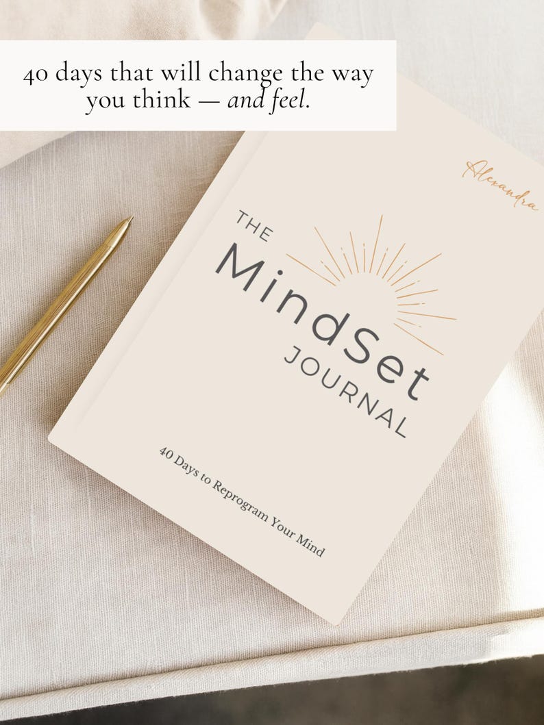 May include: A cream-coloured journal titled "The Mindset Journal" with gold lettering and a sunburst design. The text "40 days that will change the way you think - and feel" is above the journal. A gold pen rests on the surface.