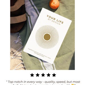 May include: A white journal titled "YOUR LIFE BY DESIGN" with gold sunburst design, resting on a green and white striped towel. A small orange fruit and a pair of brown sandals are also visible. The journal's text reads: "A guided journal for self-discovery, vision, and aligned action."
