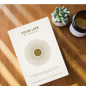 May include: A journal titled "YOUR LIFE BY DESIGN" with a sunburst design on the cover. The journal is accompanied by a small succulent in a white pot and a jar. The text reads "Not just a journal, an experience."