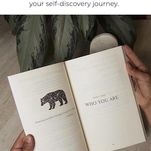 May include: An open book with the text "WHO YOU ARE" and a geometric bear illustration. The book's pages are cream-colored, and the text "Clear sections to guide you step by step in your self-discovery journey" is at the top.