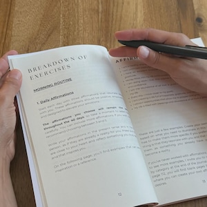 May include: An open journal with the title "BREAKDOWN OF EXERCISES" and the subheading "MORNING ROUTINE" is held open with a black pen. The page shows text about daily affirmations. The journal is on a wooden surface.