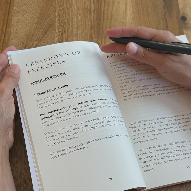 May include: An open journal with the title "BREAKDOWN OF EXERCISES" and the subheading "MORNING ROUTINE" is held open with a black pen. The page shows text about daily affirmations. The journal is on a wooden surface.