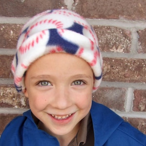 May include: A white fleece hat with a red, white, and blue baseball pattern. The hat is worn by a child with a happy expression.