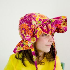 Puede incluir: Una persona joven lleva un sombrero de sol floral rosa y amarillo con un borde con volantes. El sombrero tiene lazos largos que se atan debajo de la barbilla. La persona lleva una camisa amarilla.