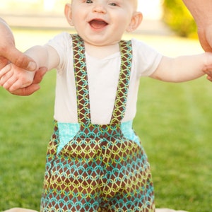 May include: A baby wearing a white shirt and brown and green patterned overalls with suspenders. The baby is smiling and holding onto an adult's hand.