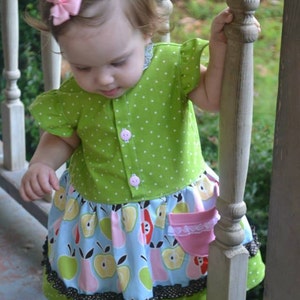 Puede incluir: Una niña pequeña con un vestido de lunares verdes y blancos con una falda de estampado de manzanas azules y verdes. El vestido tiene un lazo rosa en el pelo de la niña y un bolsillo rosa en la falda.