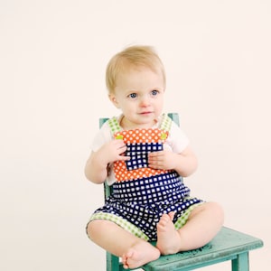 May include: A toddler wearing a blue and white gingham overall with orange and white polka dot accents sits on a teal blue wooden chair.