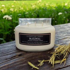 May include: A clear glass jar candle with a black label that reads "GLOWSTONE CANDLE BUZZKILL Citronella | Lemongrass | Verbena HAND-POURED IN TXI ALL-NATURAL COCONUTSOY WAX 40+ HR BURN TIME | 11OZ | 312G"
