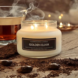 May include: A white candle in a clear glass jar with a black label that reads "GLOWSTONE CANDLE GOLDEN ELIXIR HAND-POURED IN TX | ALL-NATURAL COCONUTSOY WAX 40- HR BURN TIME | 11OZ | 312G"