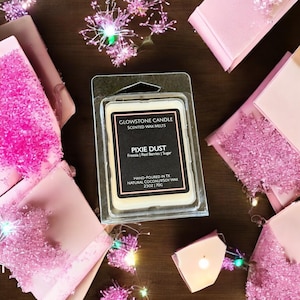 Puede incluir: Un paquete de plástico transparente que contiene un ambientador de cera perfumada Pixie Dust de Glowstone Candle. La etiqueta enumera Fresia, Bayas rojas y Azúcar como aromas. Contenedores de ambientador de cera rosa y luces de hadas rodean el paquete.