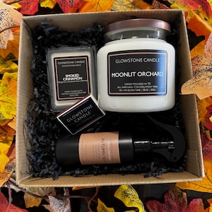 May include: A gift box containing a Moonlit Orchard candle in a glass jar, a package of Smoked Cinnamon wax melts, a small box of matches, and an Autumn Sky room spray bottle, all nestled in black paper shreds with autumn leaf decorations.