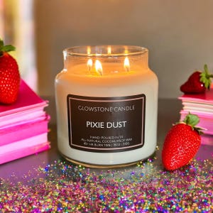 May include: A clear glass jar candle with a brown label that reads "GLOWSTONE CANDLE" and "PIXIE DUST". The label also includes the text "HAND-POURED IN TX", "ALL-NATURAL COCONUTSOY WAX", "45+ HR BURN TIME", "18OZ", and "510G". The candle is lit and surrounded by colorful glitter.