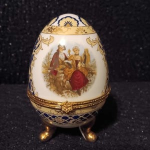 May include: An egg-shaped decorative trinket box with a hinged lid. The box is white with a painted scene of figures in period clothing. Gold trim and accents, with blue and gold patterned details. The box rests on three gold-colored feet.