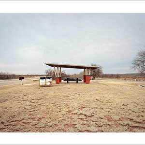May include: A covered picnic table with two trash cans sits in a grassy area with a paved road in the background. The sky is overcast and the ground is covered in dry grass.