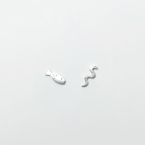 May include: Two small, silver-coloured charms. One charm is shaped like a fish with dotted markings. The other charm is a stylised snake with a curved body. Both charms are set against a white background.