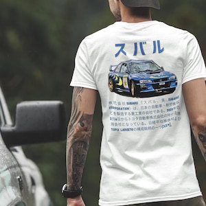 Peut inclure: Un t-shirt blanc avec une voiture de rallye Subaru Impreza bleue imprimée au dos. La voiture est entourée de texte japonais qui lit "Subaru" et comprend des informations sur la société.