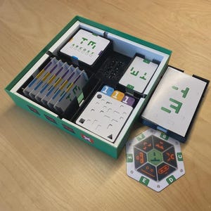 May include: A board game set in a green box with various components. The set includes cards, tiles, and a hexagonal game piece. The cards and box feature a green and white geometric design. The game is set on a wooden surface.