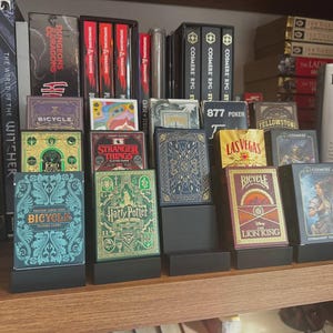 May include: Various playing card decks, including Bicycle, Harry Potter, and Disney's Lion King, are displayed on black stands. The decks have different designs and themes, with book titles visible in the background.