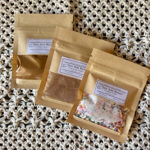 May include: Three brown paper packets with clear windows, each labeled "Just Add Butter." One contains cinnamon spice, another cocoa and cream, and the third confetti cake mix with colorful sprinkles. The packets are arranged on a textured, off-white surface.