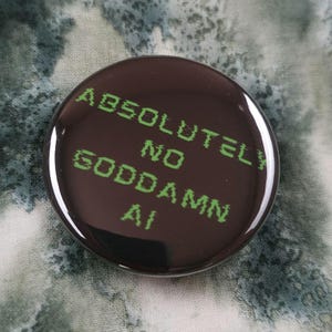 May include: A circular pin-back button with a glossy black surface. The text "ABSOLUTELY NO GODDAMN AI" is printed in a bright green, pixelated, digital-style font across the center of the button, creating a high-contrast look against the dark background.