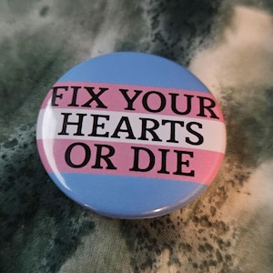 May include: A circular badge with a light blue background and the text "FIX YOUR HEARTS OR DIE" in black. The badge has a horizontal stripe in white and pink, with a shiny surface.