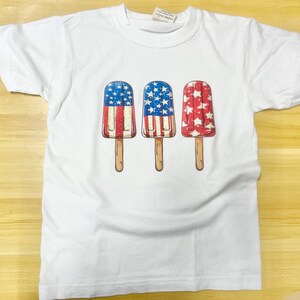 K&ouml;nnte beinhalten: Wei&szlig;es T-Shirt mit einem Motiv von drei Eis am Stiel. Jedes Eis ist mit der amerikanischen Flagge verziert.