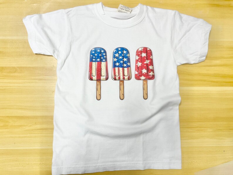 K&ouml;nnte beinhalten: Wei&szlig;es T-Shirt mit einem Motiv von drei Eis am Stiel. Jedes Eis ist mit der amerikanischen Flagge verziert.