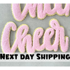 May include: Pink fuzzy letters spelling "Cheer" with gold glitter outlining. The letters are on a grey background. Next Day Shipping is written in black text below the letters.