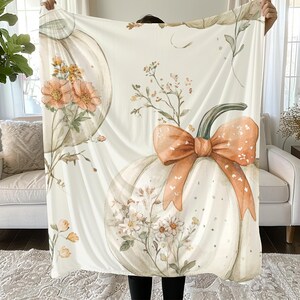 May include: Cream-colored blanket featuring a watercolor design of pumpkins, flowers, and foliage. The pumpkins have a bow and floral accents. The blanket is soft and cozy, perfect for fall decor.