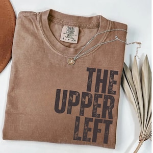 May include: A brown Comfort Colors t-shirt with the text "THE UPPER LEFT" in dark brown. A silver necklace with a small pendant is on the shirt. A brown hat is partially visible in the upper left corner.