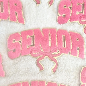 Puede incluir: Parches de fieltro rosa con letras doradas que deletrean "Senior" con un lazo rosa en cada parche.