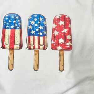 K&ouml;nnte beinhalten: Drei Eis am Stiel mit rot-wei&szlig;-blauen Designs. Die Eis am Stiel befinden sich auf einem wei&szlig;en T-Shirt.