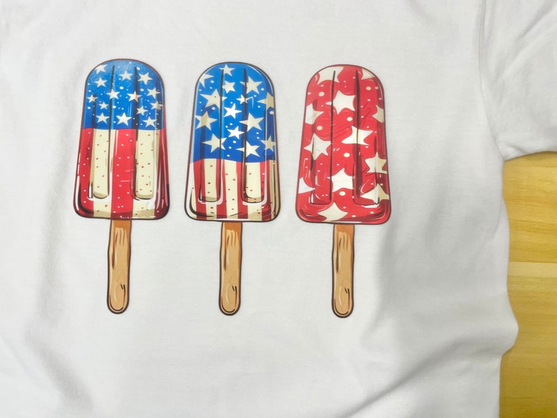 K&ouml;nnte beinhalten: Drei Eis am Stiel mit rot-wei&szlig;-blauen Designs. Die Eis am Stiel befinden sich auf einem wei&szlig;en T-Shirt.