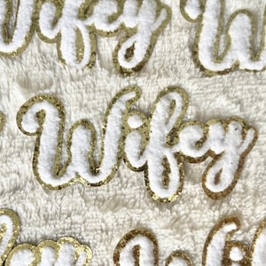 May include: A close-up of several gold glitter iron-on patches with the word "Wifey" in white felt lettering. The patches are on a white fuzzy surface.